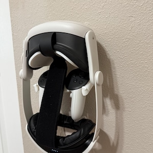 May include: A white and black VR headset with matching controllers is mounted on a wall. The headset has a black head strap and black padding around the eyes. The controllers are white with black buttons.