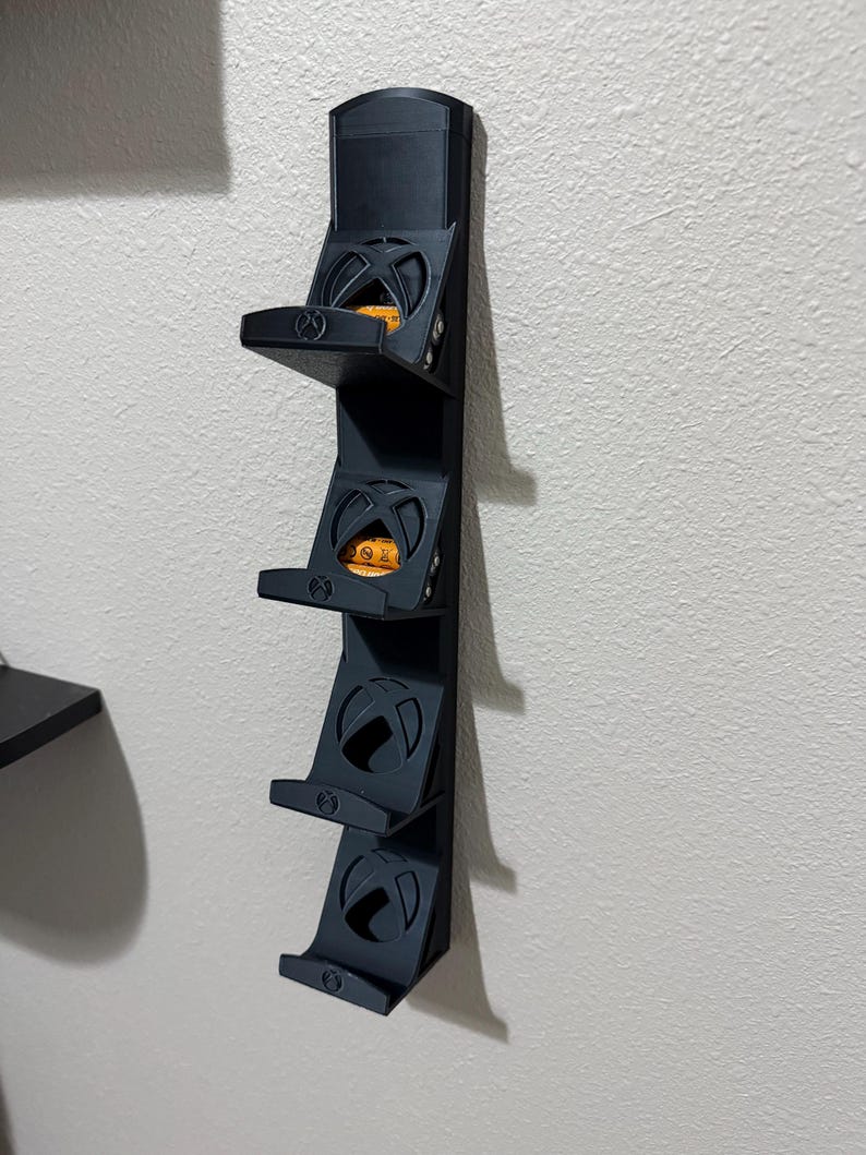 May include: A black wall-mounted storage rack with four shelves, each designed to hold a gaming console. The shelves have a circular design with an 'X' shape in the center. The rack is mounted on a white wall.