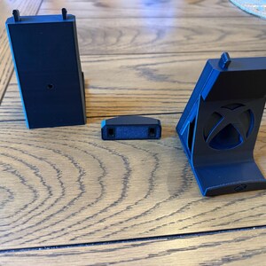 May include: Three black plastic gaming console accessories are displayed on a wooden surface. The accessories include a vertical stand, a small rectangular piece, and a charging station. The stand features the Xbox logo.