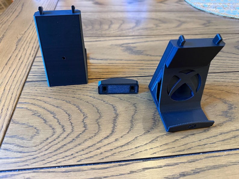 May include: Three black plastic gaming console accessories are displayed on a wooden surface. The accessories include a vertical stand, a small rectangular piece, and a charging station. The stand features the Xbox logo.
