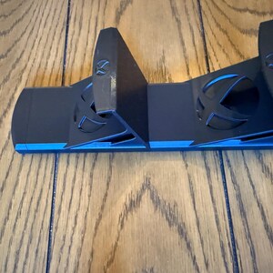 May include: Black and blue gaming console controller stand. The stand has a sleek design with the Xbox logo cut out on the front. The stand is made of plastic and has a blue accent.