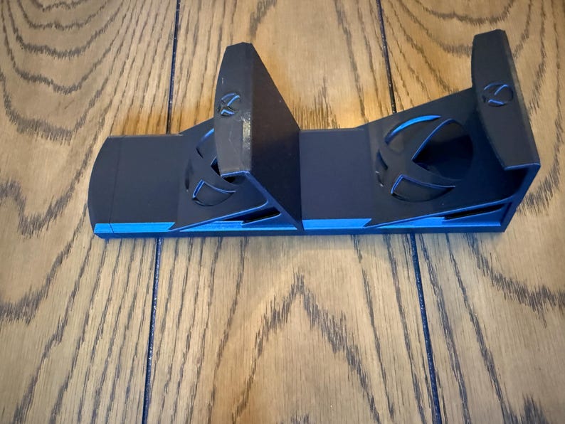 May include: Black and blue gaming console controller stand. The stand has a sleek design with the Xbox logo cut out on the front. The stand is made of plastic and has a blue accent.