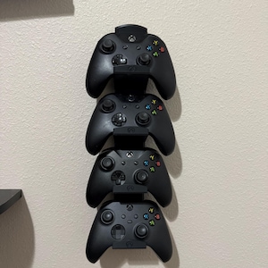 May include: Four black Xbox controllers are stacked vertically on a wall-mounted holder. Each controller has colored buttons and a central Xbox logo. The controllers are neatly arranged, suggesting a gaming setup.