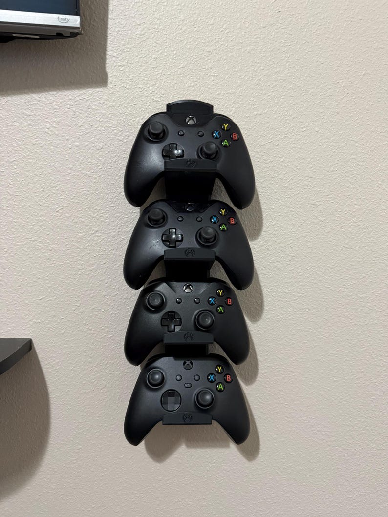 May include: Four black Xbox controllers are stacked vertically on a wall-mounted holder. Each controller has colored buttons and a central Xbox logo. The controllers are neatly arranged, suggesting a gaming setup.