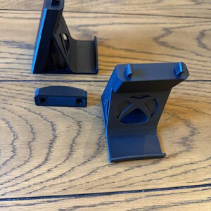 May include: Black plastic Xbox console stand with three pieces. The stand has a curved base and a vertical support with a rectangular top. The Xbox logo is visible on one of the pieces. The stand is designed to hold the console upright.