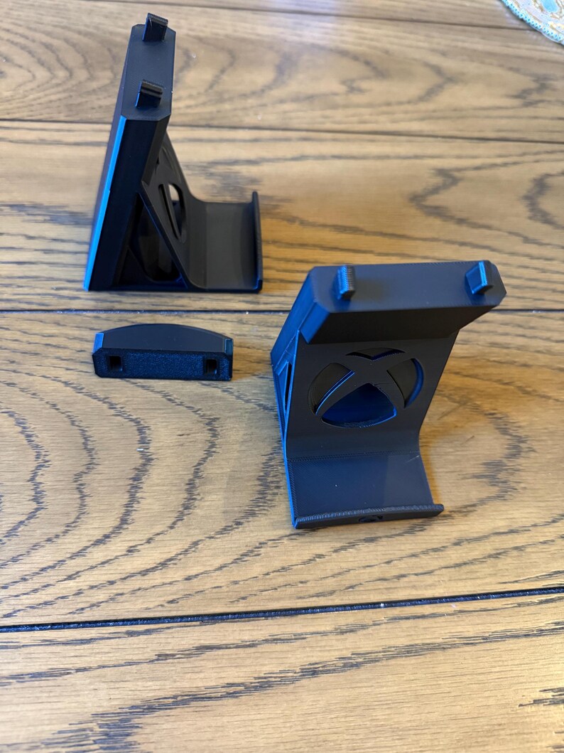 May include: Black plastic Xbox console stand with three pieces. The stand has a curved base and a vertical support with a rectangular top. The Xbox logo is visible on one of the pieces. The stand is designed to hold the console upright.