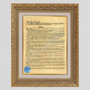 May include: A framed replica of the United States Constitution on aged parchment paper. The document features the text We The People and Article I in black ink. It is encased in an ornate gold frame with a light-coloured mat and a blue seal.