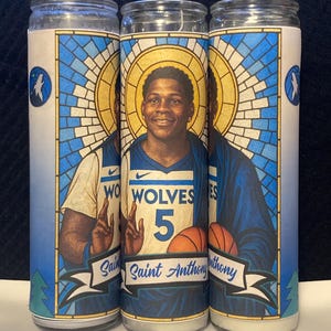 May include: Three glass prayer candles featuring a portrait of a basketball player in a blue and white jersey. The candles have a stained-glass style border in blue and white, with the words "Saint Anthony" on a banner.
