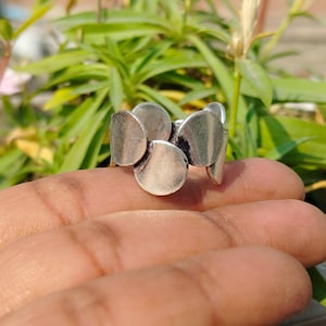 Pode incluir: Um anel de prata com um grupo de formas arredondadas sobrepostas. O anel tem um acabamento polido e é colocado sobre um fundo de folhagem verde. O design do anel é moderno e abstrato.