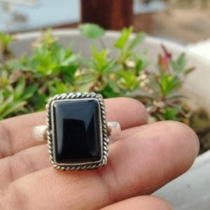 May include: A silver ring with a rectangular black stone. The stone is set in a silver frame with a rope-like design around the edges. The ring is held up by a hand.