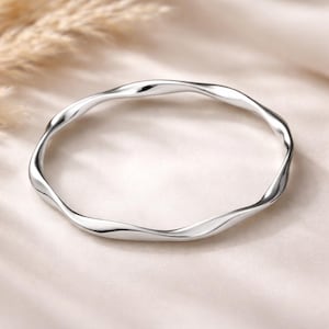 May include: A silver-coloured, wavy bangle bracelet. The bracelet has a smooth, reflective surface and a fluid, organic design. The bracelet is a simple, elegant piece of jewellery.
