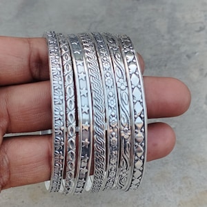 Puede incluir: Un conjunto de pulseras plateadas con intrincados diseños florales, de estrellas y geométricos. Las pulseras están apiladas, mostrando los diseños y texturas detalladas. La joyería está hecha de un material metálico brillante.