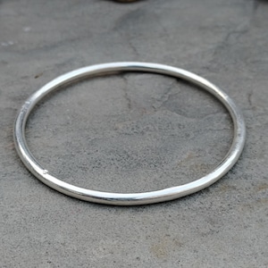 Könnte beinhalten: Ein schlichtes, silberfarbenes Armreif-Armband. Das runde Armband besteht aus glänzendem Metall und wird auf einer grauen Oberfläche gezeigt. Der Armreif ist ein klassisches Schmuckstück.