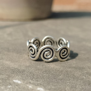 May include: A silver-colored ring featuring a repeating swirl design. The ring has a continuous pattern of open swirls, creating a decorative and eye-catching accessory. The ring is shown on a gray surface.
