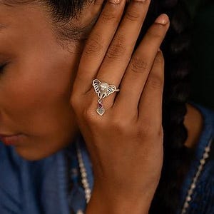 Peut inclure: Bague en argent ornée d'une tête d'éléphant et d'un pendentif en forme de cœur. La bague comporte une petite pierre précieuse rouge et une pierre nacrée. La bague est portée au doigt.