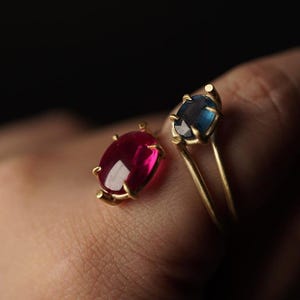 May include: Two gold rings, one with a ruby-red oval gemstone and the other with a blue oval gemstone. The rings are set against a dark background, highlighting the vibrant colours of the gems.