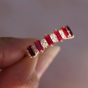 Peut inclure: Une bague en or ornée de pierres rouges rectangulaires alternées et de petites pierres claires rondes. La bague présente une série de pierres rouges vibrantes, entrecoupées de pierres claires scintillantes, créant un jeu de couleurs et de lumière.