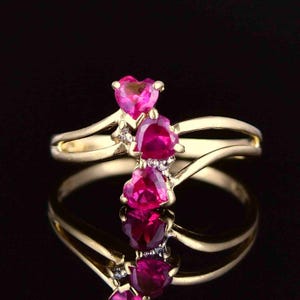 Puede incluir: Un anillo de oro con tres piedras preciosas de color rosa vibrante en forma de corazón dispuestas en un grupo vertical. La banda tiene un acabado liso y pulido y un diseño dividido que se curva alrededor de las piedras centrales, acentuadas por pequeñas piedras claras.
