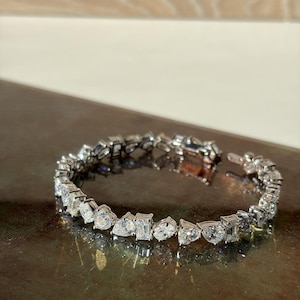 Può includere: Un bracciale tennis color argento caratterizzato da un motivo ripetuto di pietre trasparenti sfaccettate alternate con taglio a goccia, smeraldo e rotondo. Il bracciale è realizzato con una catena a maglie metalliche flessibili e una chiusura.