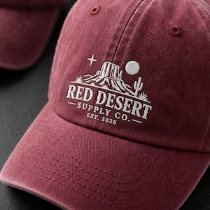 Può includere: Un cappellino da baseball color marrone scuro con un ricamo bianco raffigurante un paesaggio desertico, una stella e una luna. Il testo recita RED DESERT SUPPLY CO. EST. 2026. Il cappellino presenta una visiera curva e occhielli cuciti sulla calotta.
