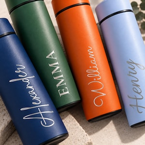 May include: Four cylindrical metal water bottles in navy blue, forest green, burnt orange, and light blue. Each bottle features a black cap and a personalised name engraved in a silver script or block font: Alexander, EMMA, William, and Henry.