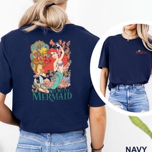 May include: A navy blue short-sleeve t-shirt featuring a large graphic on the back of Ariel and Flounder from The Little Mermaid with floral accents and the text The Little Mermaid. The front chest area displays a small The Little Mermaid logo.