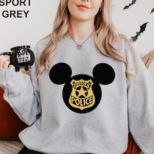 May include: Sport grey sweatshirt featuring a black Mickey Mouse silhouette with a Zootopia Police badge design in gold and black. The sweatshirt has a crew neck and long sleeves. A black mug with the text "Witches Brew" is held in the left hand.