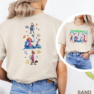 May include: A sand-coloured t-shirt with a colourful graphic of characters from Zootopia 2. The back of the shirt displays a vertical arrangement of Zootopia characters and icons. The front of the shirt has the text "Zootopia 2" in green and blue.