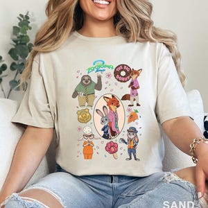 K&ouml;nnte beinhalten: Ein sandfarbenes T-Shirt mit einem Zootopia 2 Grafikdruck. Das Design zeigt Charaktere wie Nick Wilde, Judy Hopps, Flash und Bellwether sowie Symbole wie einen Donut, eine Karotte, ein Abzeichen und ein pfotenf&ouml;rmiges Eis am Stiel.