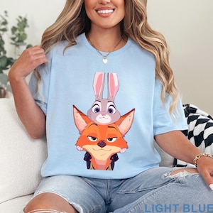 May include: Light blue t-shirt featuring a graphic of the Zootopia characters Judy Hopps and Nick Wilde. The text "LIGHT BLUE" is printed at the bottom. The shirt is a light blue color.