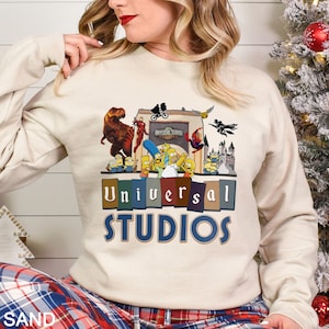 May include: A sand-colored sweatshirt featuring a colorful graphic of Universal Studios characters and the park's logo. The design includes dinosaurs, superheroes, and cartoon characters. The text "Universal Studios" is displayed prominently.