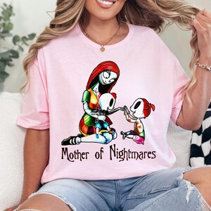 Puede incluir: Camiseta rosa claro con un gr&aacute;fico de un personaje con cabello rojo y apariencia cosida, con dos personajes m&aacute;s peque&ntilde;os. El texto "Mother of Nightmares" est&aacute; impreso debajo. La camiseta es de material suave.