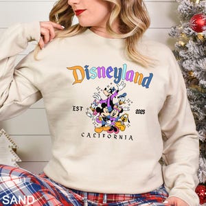 Puede incluir: Una sudadera de color arena con el logotipo de Disneyland en naranja y azul, con un gr&aacute;fico de Mickey Mouse, Minnie Mouse, Donald Duck, Goofy y Pluto. El texto "EST 2025 CALIFORNIA" tambi&eacute;n est&aacute; impreso.
