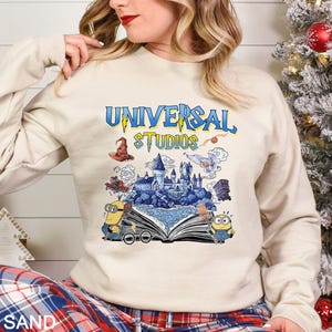 Op de afbeelding: Zandkleurige sweatshirt met een kleurrijke afbeelding van het Universal Studios-logo en themaparkelementen. Het ontwerp bevat een tovenaars hoed, een kasteel en Minions. De tekst "UNIVERSAL STUDIOS" is in blauw en geel.