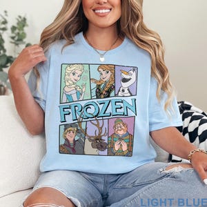 May include: Light blue t-shirt featuring the word "FROZEN" and images of characters from the animated movie. The shirt has a crew neck and short sleeves. The text "LIGHT BLUE" is printed at the bottom of the shirt.