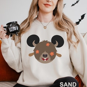 May include: Sand-colored sweatshirt featuring a cartoon bear face with large black ears, brown fur, and orange flower accents. The bear has a smiling expression with two white teeth. The word "SAND" is printed at the bottom.