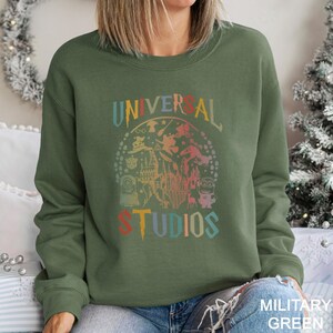 May include: Olive green sweatshirt with the words "UNIVERSAL STUDIOS" in a colorful, vintage-style design. The graphic includes various characters and imagery associated with Universal Studios.