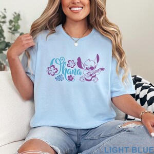 Op de afbeelding: Een lichtblauw T-shirt met korte mouwen en een paarse afbeelding van het personage Stitch die op een ukelele speelt, vergezeld van de tekst Ohana en diverse illustraties van hibiscusbloemen op het midden van de borst.