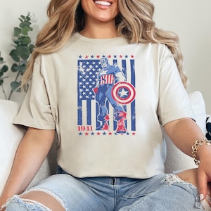 May include: Beige t-shirt featuring a distressed graphic of Captain America with his shield, set against a backdrop of the American flag. The year "1941" is printed below the image. The design is in shades of blue, red, and white.