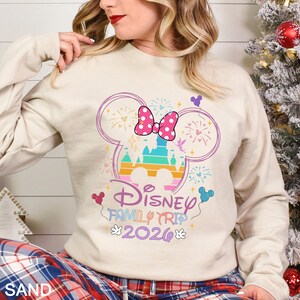 May include: A sand-colored sweatshirt featuring a colorful Disney castle design within a Mickey Mouse head outline. The design includes a pink bow, fireworks, and the text "Disney Family Trip 2026".