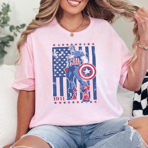 May include: Pink t-shirt featuring a graphic of Captain America in a patriotic design. The design includes the American flag, stars, and the year 1941. The superhero is holding his shield. The shirt is a casual, round-neck style.