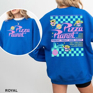 Pu&ograve; includere: Una felpa girocollo blu reale con una grafica di Pizza Planet. La parte anteriore mostra il logo, mentre il retro presenta un design dettagliato con alieni, un motivo a scacchi e testo che include Interstellar Fast-Food Fueling Station.