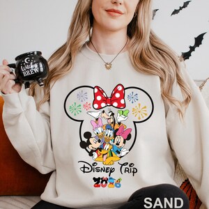 May include: Sand-colored sweatshirt featuring a large graphic of Disney characters, including Mickey and Minnie Mouse, Donald and Daisy Duck, and Goofy. The design includes a red bow with white polka dots and the text "Disney Trip 2023".