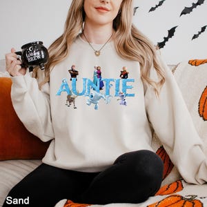 K&ouml;nnte beinhalten: Ein sandfarbenes Sweatshirt mit Rundhalsausschnitt, das das Wort AUNTIE in einer blauen, eisigen Schriftart zeigt, verziert mit verschiedenen animierten Charakteren aus der Frozen-Filmreihe. Das Sweatshirt wird mit einem schwarzen Kesselbecher kombiniert.