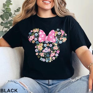 May include: Black t-shirt featuring a floral Minnie Mouse design. The design includes a pink polka-dot bow and a floral arrangement in the shape of Minnie's head. The word "BLACK" is printed at the bottom.