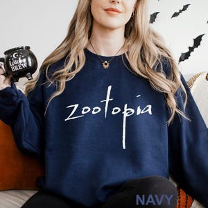 May include: Navy blue sweatshirt with the word "zootopia" in white script. The word "NAVY" is printed in white at the bottom. A black mug with the words "Witch's Brew" is held in the left hand.