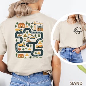 May include: A sand-colored t-shirt featuring a back graphic of a construction site with roads, trucks, a crane, houses, and warning signs. The front chest area displays the black text "TIRED DADS CLUB" in a circular, cursive-style font design.