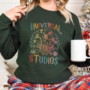 Pode incluir: Camisola verde floresta com um gr&aacute;fico colorido "UNIVERSAL STUDIOS" com personagens de parques tem&aacute;ticos. A camisola &eacute; de gola redonda e tem mangas compridas. Uma caneca branca &eacute; segurada na m&atilde;o.