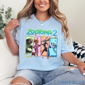 May include: Light blue t-shirt featuring the text "ZOOTOPIA 2" above four character panels. The panels display cartoon images of a sloth, fox, rabbit, and snake. The shirt is light blue and the words "LIGHT BLUE" are printed at the bottom.