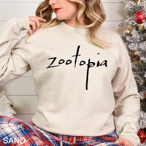 May include: A sand-colored sweatshirt with the word "Zootopia" in black script. The long-sleeved crew neck top is worn with plaid pajama pants. A Christmas tree with red ornaments is in the background.